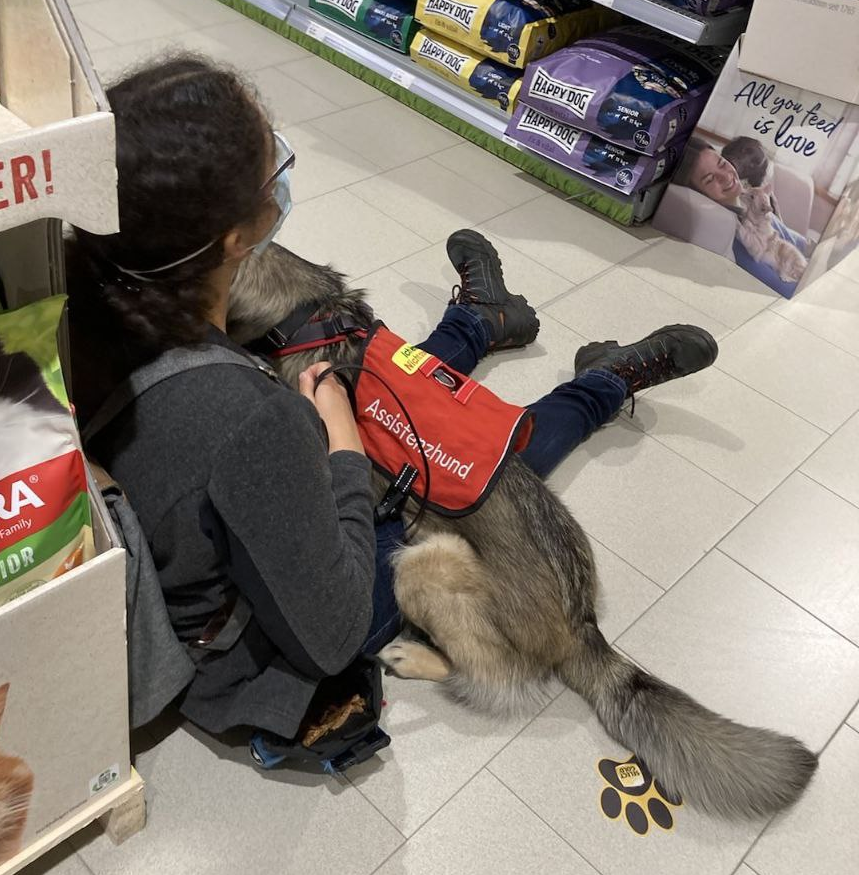 Frau sitzt mit ausgestreckten Beinen am Boden, Hund liegt quer über die Beine ausgestreckt.