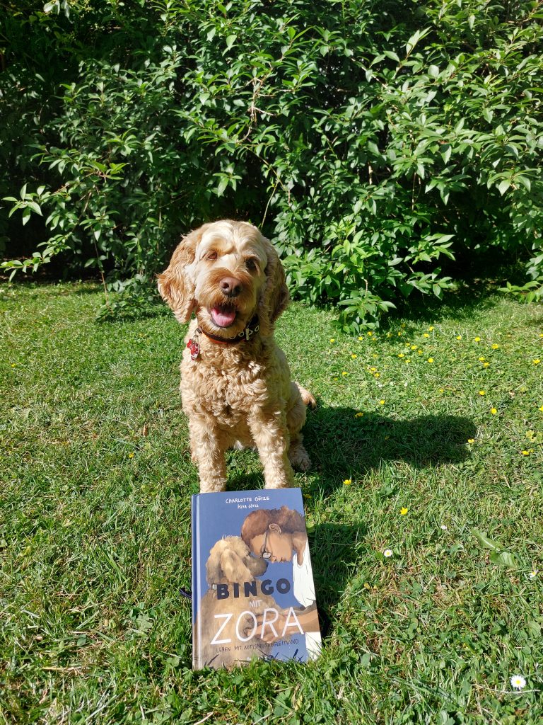 Hund Zora sitzt auf einer Wiese vor einem Busch. Vor ihr liegt das Buch "Bingo mit Zora".