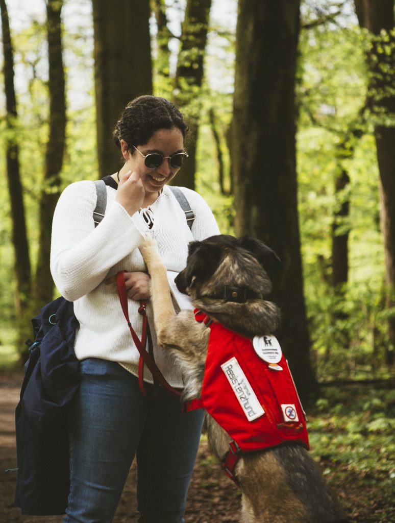Nora steht in einem Wald. Hund Mia mit Kenndecke springt an ihr hoch. Mias Pfoten erreichen Noras Brust. Die beiden schauen sich an.